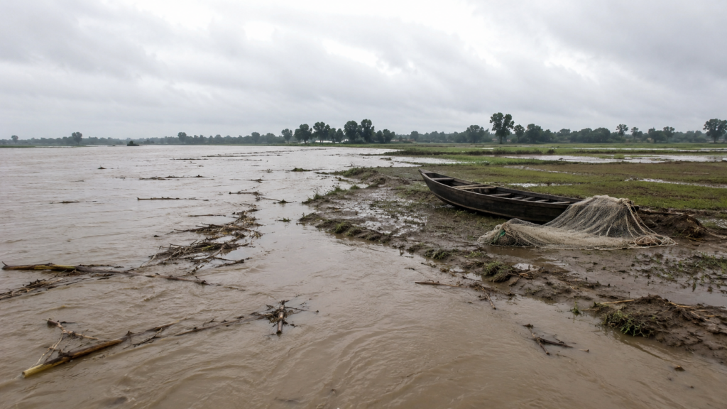 Muddy floodwater covering farmland in north Bihar, with a fishing boat and unused net on the bank.